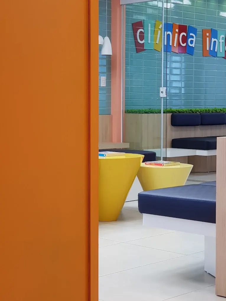 Recepção da clínica com porta laranja, mesas amarelas e bancos azul-marinho